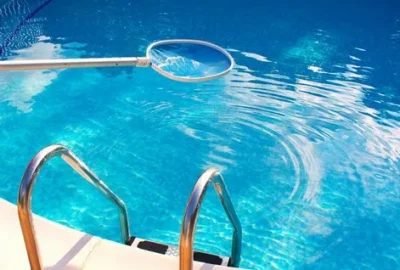 A pool skimmer net floats on the surface of clear blue swimming pool water near metal ladder rails.