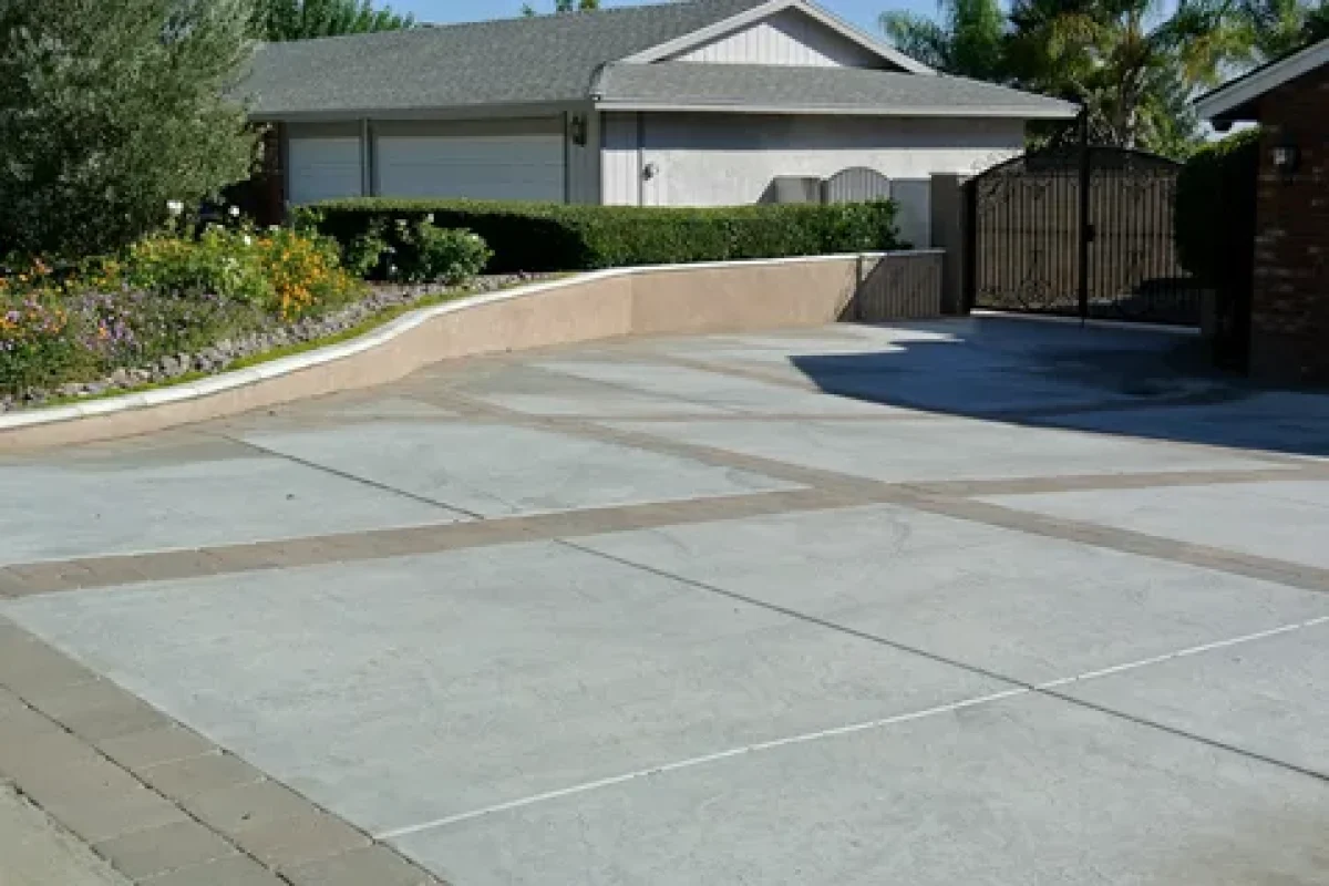 Concrete driveway with decorative paver borders leading to a house and a black wrought iron gate.