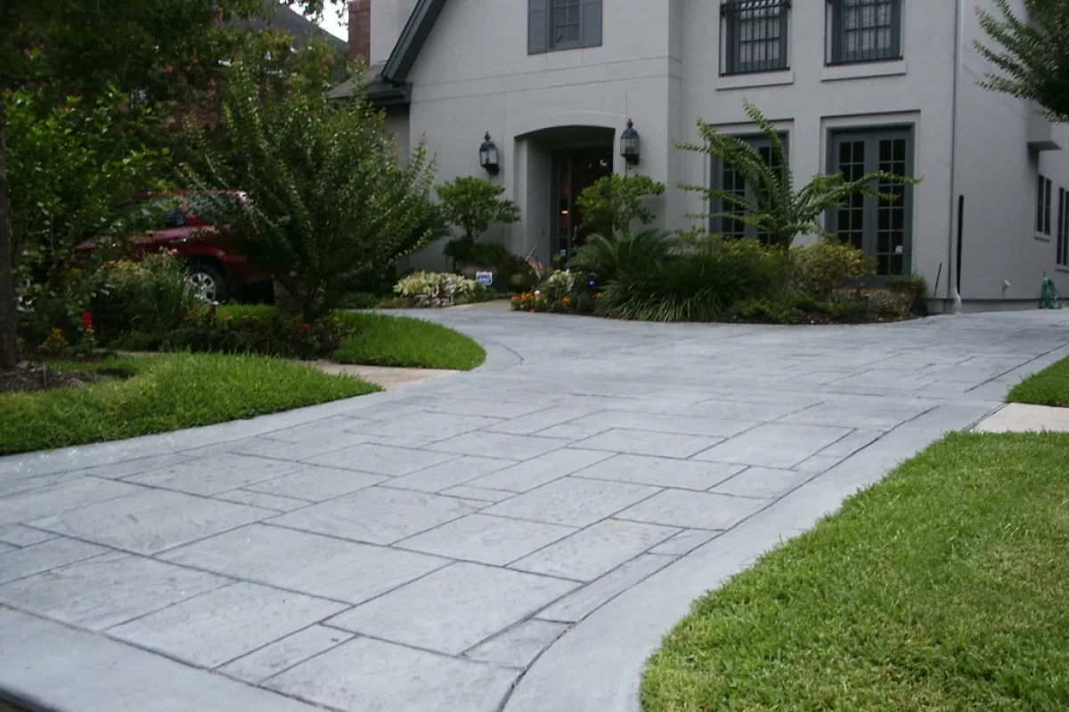 Gray stamped concrete driveway with a stone tile pattern leading to a light-colored house with green landscaping.