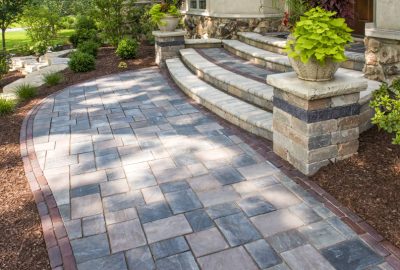 Curved stone paver walkway and steps leading to a house entrance, surrounded by mulch and lush greenery.