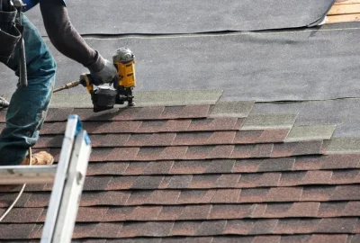 Roofer using a pneumatic nail gun to install brown asphalt shingles on a roof.