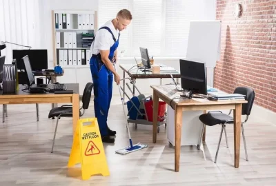 A janitor in blue overalls mops a floor in a modern office next to a yellow caution wet floor sign.