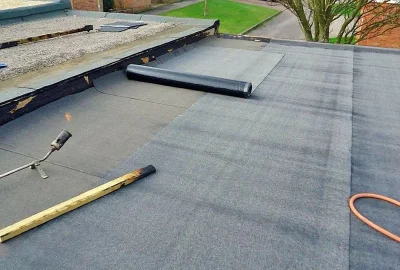 Torch-on roofing membrane being installed on a flat roof with a propane torch and wooden tamper.
