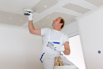 A man in white overalls uses a putty knife to apply joint compound to a ceiling.