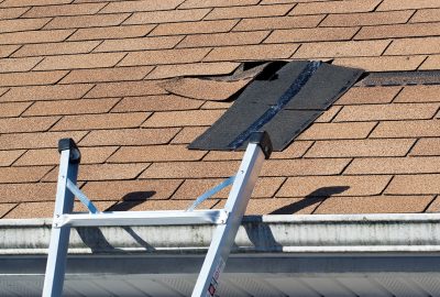 Silver ladder leaning against a brown shingle roof with a damaged area exposing black underlayment.