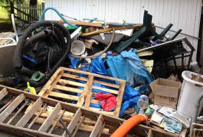 A large pile of cluttered junk, including wooden pallets, a ladder, blue tarps, a black hose, and various household items.