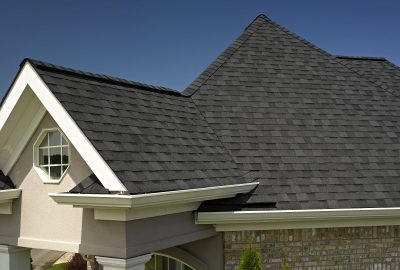 Dark grey asphalt shingles on a residential roof with white gutters and trim under a clear blue sky.