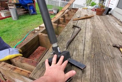 A person's hand stabilizes a black hydraulic jack positioned on a wooden deck during a construction project.