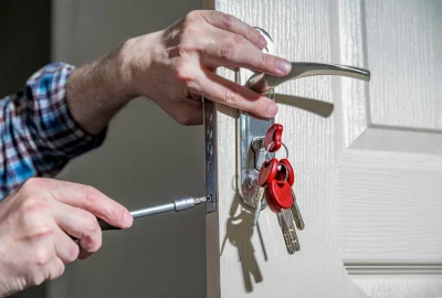 Person repairing a white door handle with a screwdriver, keys with red tags hanging from the lock.