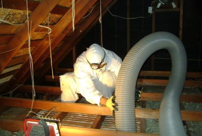 Worker in full protective gear working with a large grey flexible duct in a dark attic.