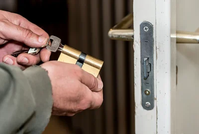 Hands holding a brass door lock cylinder and key in front of a white door.