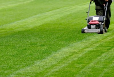 A person pushes a lawnmower across a wide, manicured green field, creating striped patterns in the grass.