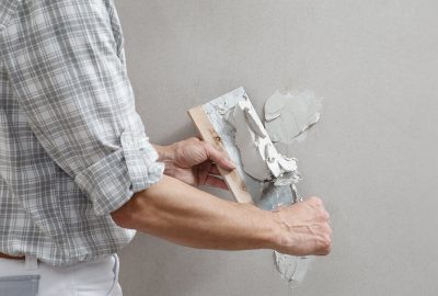 A worker wearing a plaid shirt uses a trowel to apply joint compound to a drywall seam.