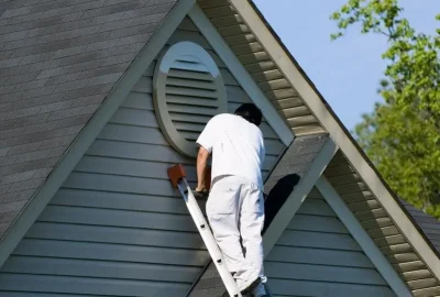 A painter in white clothing on a ladder painting the trim of a circular gable vent on a house.