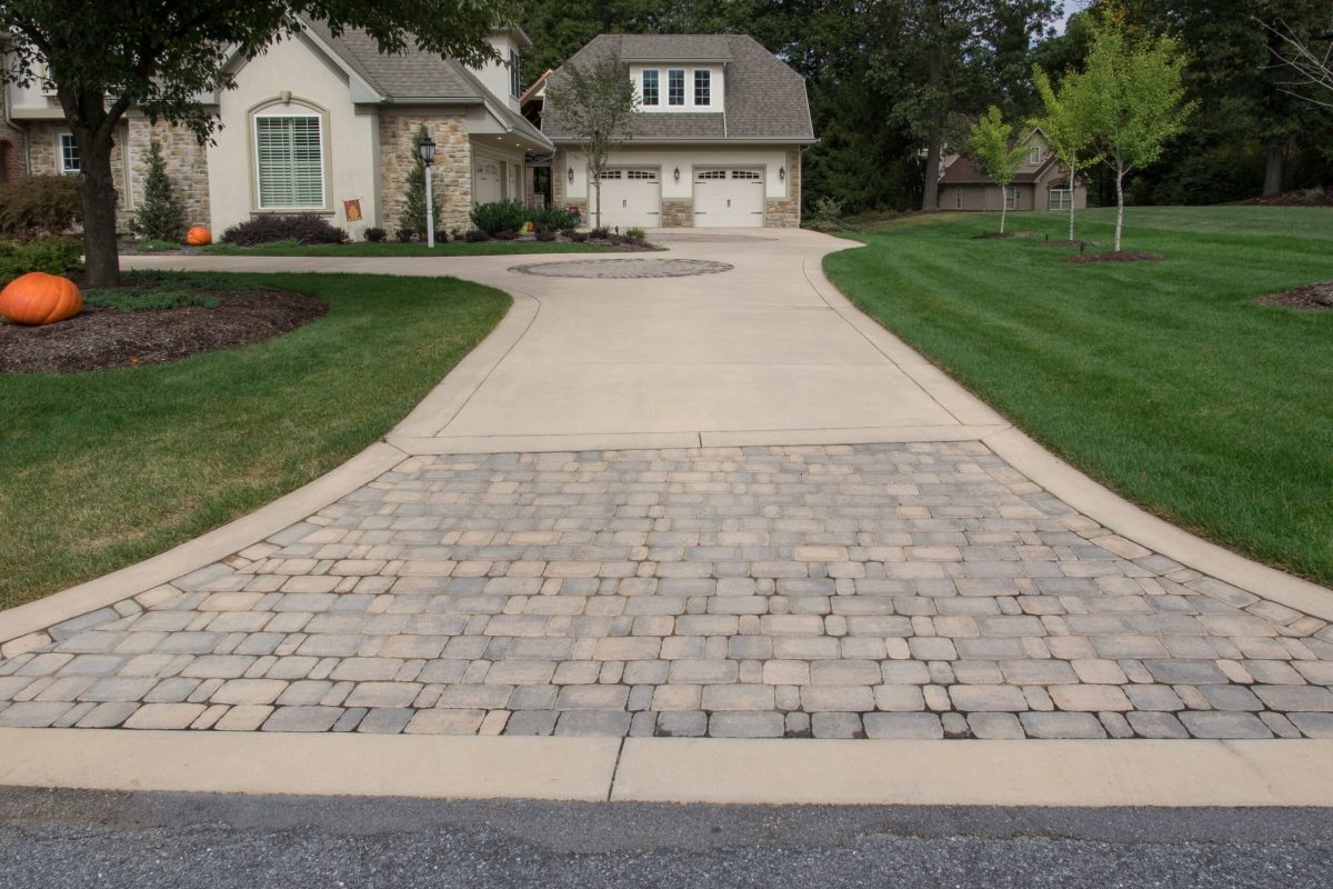 Concrete driveway with a cobblestone entrance leading to a large house with a two-car garage and manicured lawn.