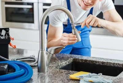 A plumber in blue overalls uses a wrench to repair a modern kitchen faucet.