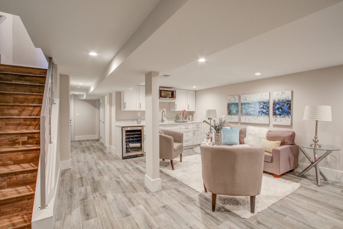 Modern finished basement with a small white kitchenette and a sitting area featuring a pink sofa and neutral armchairs.