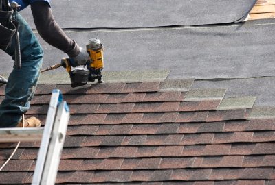 Roofer installing brown asphalt shingles on a roof using a pneumatic nail gun.