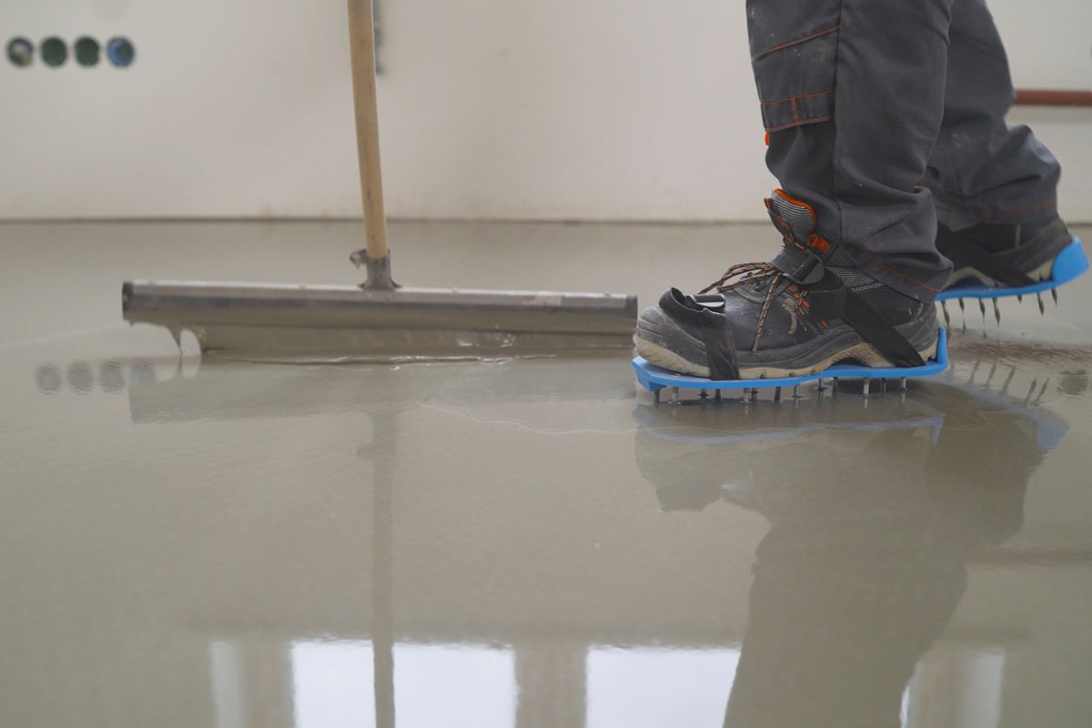 Worker in blue spiked shoes walking on wet concrete next to a concrete bull float.