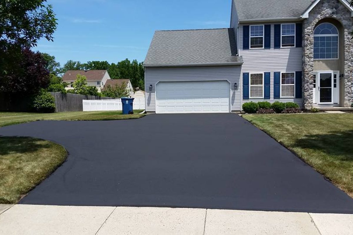 Freshly paved black asphalt driveway leading to a grey suburban house with blue shutters.