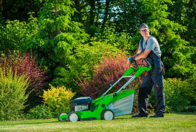 A man in overalls and safety glasses stands behind a bright green lawn mower in a lush, green garden.