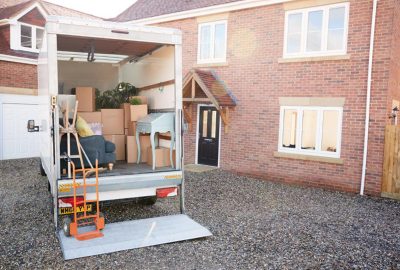 A moving truck with an open back filled with boxes, a blue sofa, and a desk parked in a gravel driveway by a brick house.
