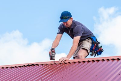 Roofer using a power drill on a red corrugated metal roof under a blue sky.