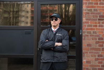 Security guard in a black jacket, cap, and sunglasses standing with arms crossed in front of a glass door.
