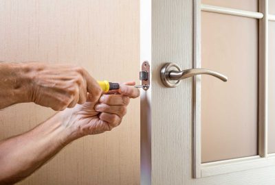 Close-up of hands using a yellow-handled screwdriver to adjust a metal door latch.