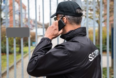 Security guard in a black jacket and cap talking on a smartphone while standing by a metal fence.