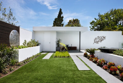 Modern minimalist courtyard with green lawn, geometric concrete paths, white walls, and artistic metal sculptures.