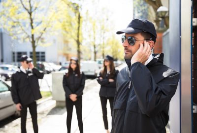 Security guard in a black uniform, cap, and sunglasses touches his earpiece, with other guards blurred in the background.