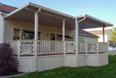 Large covered beige deck with a white roof structure attached to a house, overlooking a green lawn.