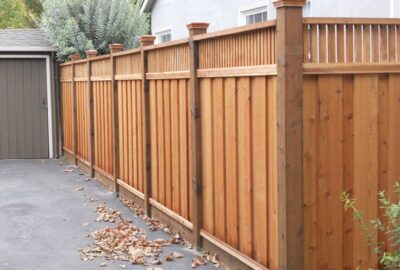 A long, light-brown wooden privacy fence with vertical slats and decorative post caps along a paved driveway.