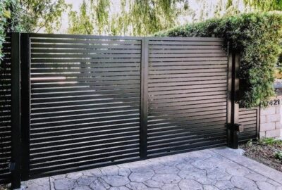 Modern black horizontal slat metal gate on a stone pavement with greenery in the background.