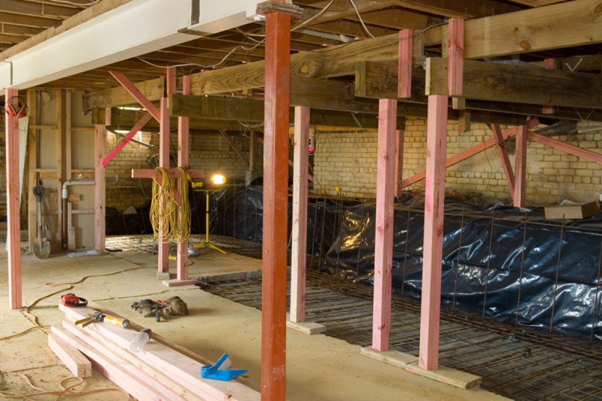 Basement under construction with red support posts, overhead wooden beams, and rebar on the floor.