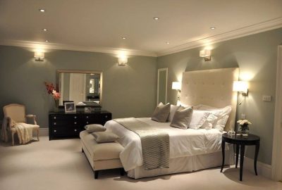 Modern bedroom with a large white bed, light grey walls, a black dresser with a mirror, and a cream armchair.