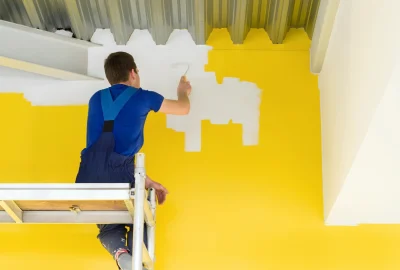 A man in blue overalls on a scaffold paints a bright yellow wall with white paint.