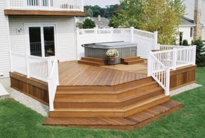 Multi-level wooden deck with white railings and a hot tub, attached to a white house and overlooking a green lawn.
