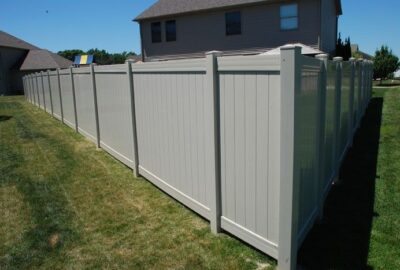 A long, light gray vinyl privacy fence runs along a green lawn next to a house under a clear blue sky.