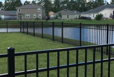 Black metal fence enclosing a green lawn and pond in a residential neighborhood.