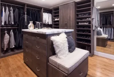 A large walk-in closet with dark wood cabinetry, a marble-topped island, a built-in bench with pillows, and hanging clothes.