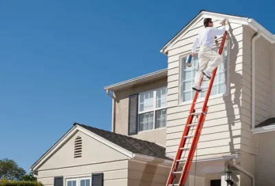 *   Image content: A man in white clothes is on a tall orange ladder, painting the upper trim/fascia of a beige house.
    *