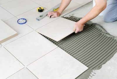 Hands placing a large square floor tile onto combed adhesive during a tiling installation.