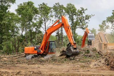 Orange excavators clearing debris and branches in a wooded area near a wood chipper.
