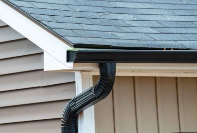 Black gutter and corrugated downspout on a house with grey shingles and tan siding.