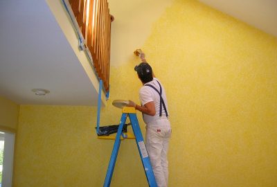 A man on a ladder uses a sponge to apply a yellow faux-finish paint texture to a high wall.