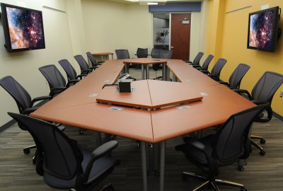 Empty conference room with a large U-shaped wooden table and black mesh chairs. Two monitors show nebula patterns.