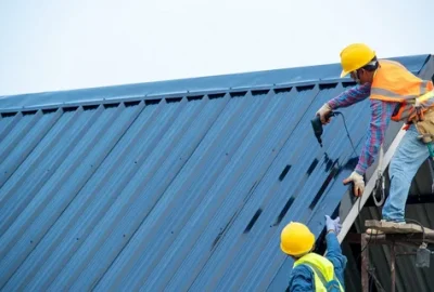 Two construction workers in hard hats and safety vests installing a blue corrugated metal roof.