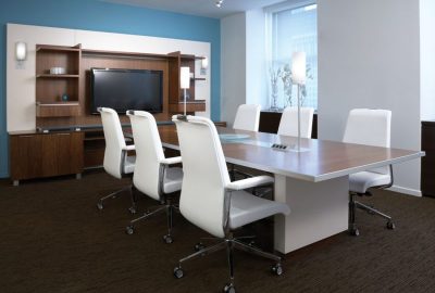 Modern conference room with a large wooden table, six white ergonomic chairs, and a wall unit with a TV.